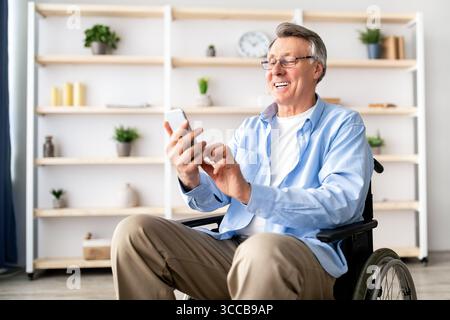 Happy disabled older man in wheelchair using cellphone, browsing web or watching movie at retirement home Stock Photo