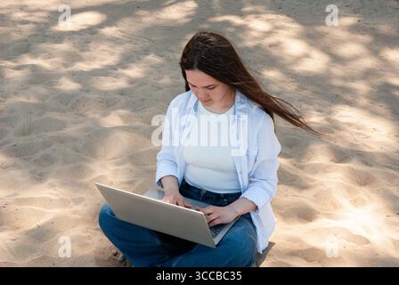 Young woman with long brown hair sitting cross legged on sandy ground in a sunny park, working on a laptop, symbolizing remote work, freelancing, and Stock Photo