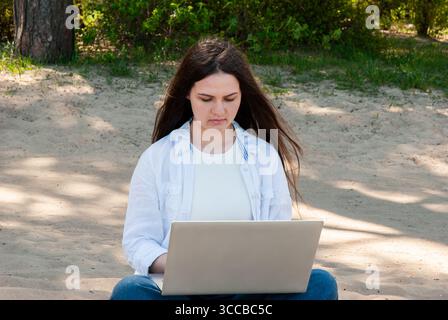 Young woman with long brown hair sitting on sandy ground in a sunny park, working on a laptop, symbolizing remote work, freelancing, and digital nomad Stock Photo