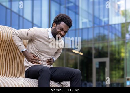 A man dressed in business casual attire experiences discomfort, holding his stomach, while seated outdoors. Stock Photo