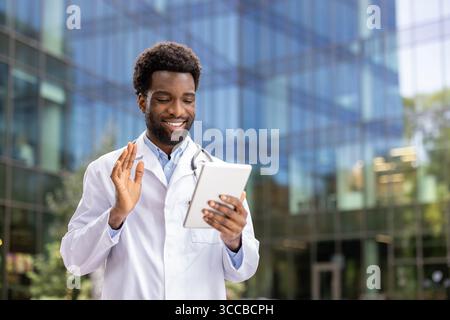 A cheerful doctor in a white coat engages in a video call using a tablet computer, standing outside glass facade, symbolizing modern healthcare commun Stock Photo