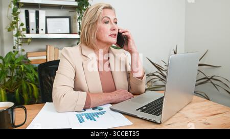 Senior Businesswoman Talking on Phone While Working on Laptop at Office Desk Stock Photo