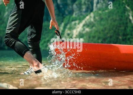 A person steps out of a bright red kayak, splashing water as they reach the shore of a serene mountain lake surrounded by lush green hills. Stock Photo
