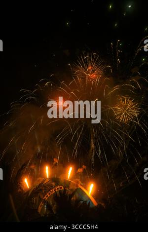 Golden fireworks burst above a dome stage with vertical fire jets and palm silhouettes, set against a dark night sky Stock Photo