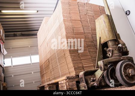 A large stack of neatly arranged cardboard boxes sits on a wooden pallet inside a warehouse. Natural light enters through the open door, illuminating Stock Photo