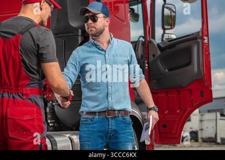Two men engaged in a professional handshake outside a red truck, showcasing collaboration in the transportation industry against a cloudy background. Stock Photo