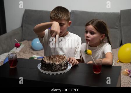 Two excited children sit together at a low table, ready to enjoy their cake. Vibrant balloons surround them while they share the joy of a delightful b Stock Photo