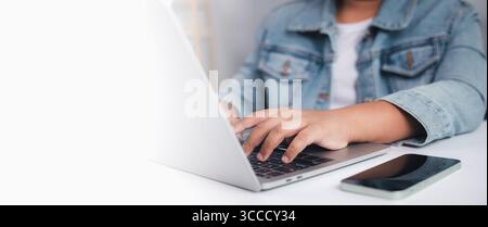 Person typing on laptop with smartphone placed on desk, copy space on left, symbolizing remote work, digital communication, and the use of technology Stock Photo