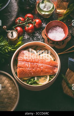 Raw salmon fillet in bamboo steamer on dark rustic kitchen table with fresh ingredients and tools. Healthy eating and cooking. Dieting concept. Asian Stock Photo