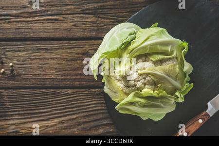 Close up of whole raw cauliflower on black cutting board with knife at rustic wooden kitchen table. Healthy uncooked garden vegetable. Cooking prepara Stock Photo