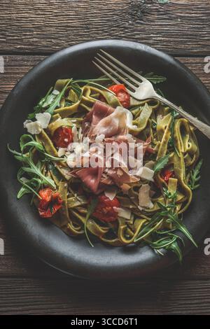 Close up of pasta dish with noodles, arugula, tomatoes, ham and cheese in black bowl with fork on rustic wooden kitchen table. Traditional Italian foo Stock Photo