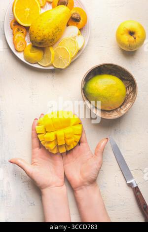 Mango preparation step by step. Female hands holding half diced mango on white background with coconut bowl and fruits plate, top view. Stock Photo