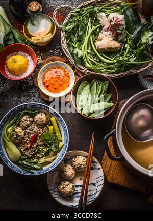 Asian food. Tasty noodle soup with vegetables and meat balls in bowl topped with sesame seeds and chili on rustic kitchen table with ingredients: herb Stock Photo