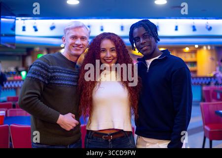 Three young adult friends smiling together inside a bowling alley, enjoying their free time together Stock Photo