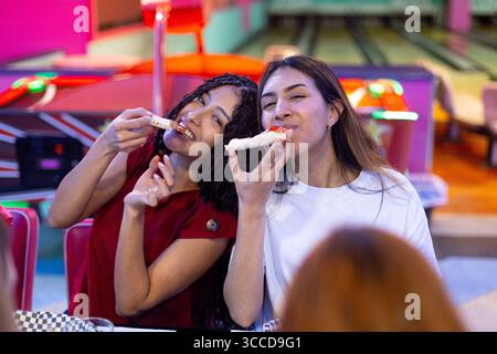 Two female friends enjoying pizza slices at a bowling alley restaurant, having fun and sharing a meal together Stock Photo