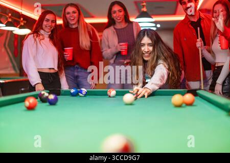 Young woman takes her shot in a game of billiards, surrounded by her friends cheering her on in a lively bar setting Stock Photo