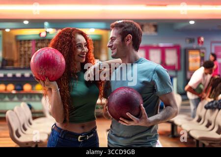 Young couple holding bowling balls, smiling joyfully at each other while enjoying a fun day at the bowling alley together Stock Photo