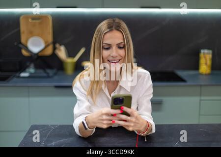 Happy businesswoman using mobile phone in modern kitchen, enjoying break time and connecting with friends and family Stock Photo