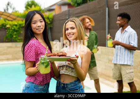 Two women enjoying grilled food and refreshing lemonade are celebrating at a lively poolside barbecue with friends on a sunny summer day Stock Photo
