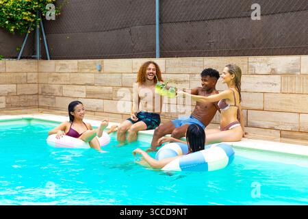 Group of cheerful multi ethnic friends toasting and drinking cocktails, sitting at the edge of a swimming pool during a summer party Stock Photo