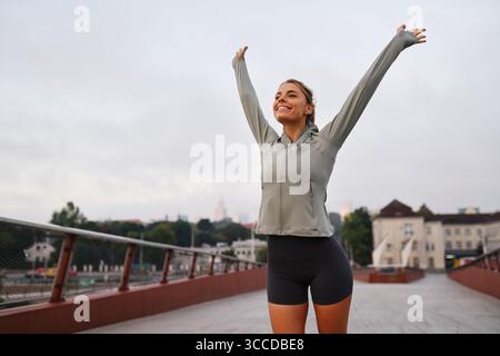A fit and energetic young woman is engaging in a stretching exercise outdoors, smiling cheerfully against a backdrop of a cloudy sky, promoting the im Stock Photo