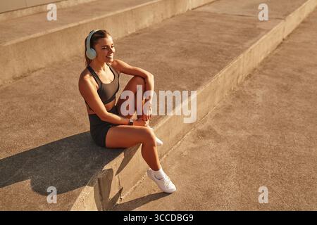 A young woman is sitting comfortably on a set of concrete steps, wearing headphones, fully absorbed in her favorite music, surrounded by a peaceful an Stock Photo