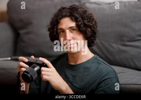 Man with curly hair holding camera, sitting on gray sofa Stock Photo
