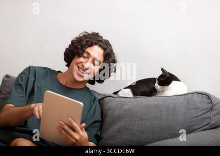 Smiling man using tablet on sofa with a resting cat beside him Stock Photo