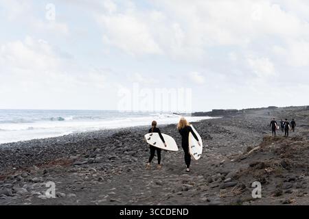 Two women surfers carrying surfboards walking on black volcanic sand beach towards the ocean Stock Photo
