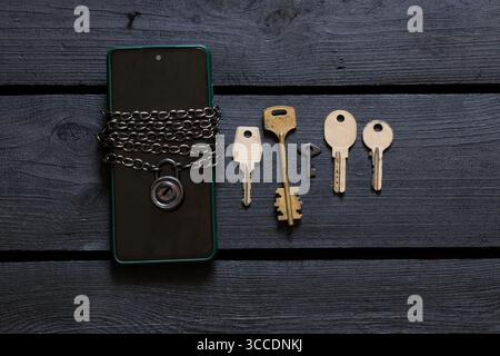 Smartphone in a green case wrapped with a metal chain with a padlock, five different keys on a black wooden surface Stock Photo