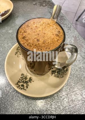 A frothy hot traditional hailam coffee in a transparent glass cup with spoon, on decorative saucer in a rustic kopitiam cafe kuala lumpur malaysia Stock Photo