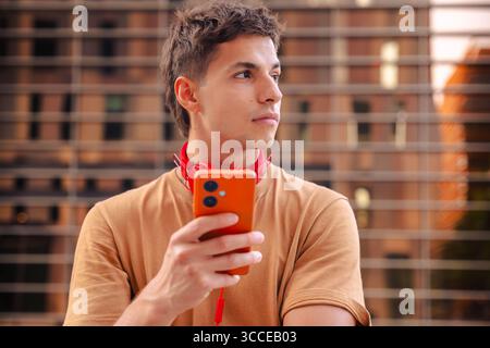 Young man holding smartphone and looking away with thoughtful expression outdoors Stock Photo