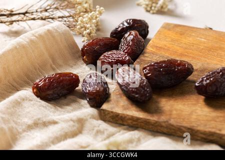 Fresh Medjool dates on a wooden board with linen cloth and dried flowers, highlighting their dark brown color, natural wrinkles, and rich texture. Per Stock Photo