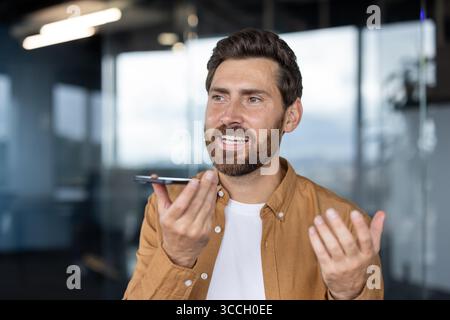 A professional man interacts with a smartphone, using its voice command feature, in an office environment with contemporary decor and natural lighting. Stock Photo