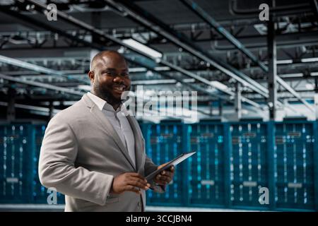 Happy technician in server hub checking recovery plan on tablet, monitoring energy consumption. Smiling data center employee using device, making sure sensors are functioning optimally Stock Photo