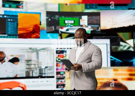 Smiling person uses tablet, streaming online video content during leisure time in modern digital environment. Joyful man browses social media app in front of digital backdrop displaying clips collage Stock Photo