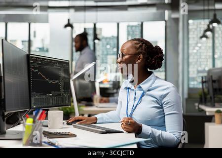 Day trader working for hedge fund company buys and sells stocks to capitalize on market fluctuations. African american woman in prop firm office uses price charts to identify fast profit opportunities Stock Photo