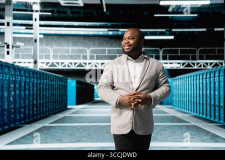Smiling admin walking in server room, overseeing mainframes gear used for managing databases. Jolly IT specialist in data center workplace doing checkup on rackmounts storing information Stock Photo