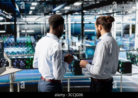 Data scientists team industrial platform analyzing performance outputs with laptop on server hub. Data center IT professionals managing server rigs configurations for AI processing. Stock Photo