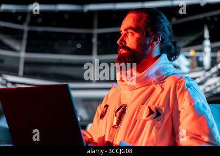 Astronaut works on notebook to troubleshoot red light warning triggered by system error in orbital station. Spaceship crew member using device to investigate malfunction affecting operations Stock Photo