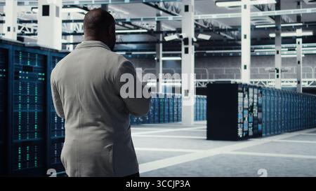 Engineer walking in server farm, automating system diagnostics using machine learning predictions. IT specialist working in data center using notebook to configure gear for AI tasks, camera A Stock Photo