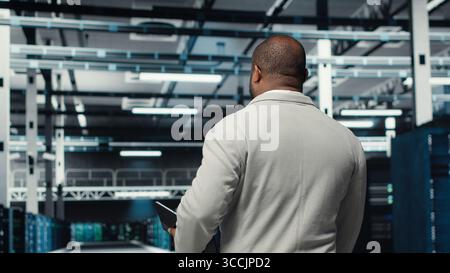 Admin walking in high tech data center facility with multiple rows of fully operational supercomputers. African american man overseeing server hub built for digital storage systems, camera B Stock Photo