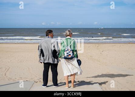 Egmond An Zee, Netherlands - 27 Aug 2024: An elderly couple stands on a sandy beach facing the sea under a sunny sky with wind turbines and a ship vis Stock Photo
