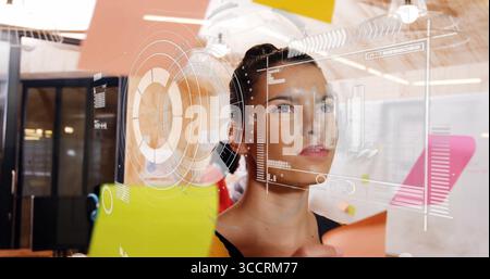 Woman in black sleeveless top arranging sticky notes on glass partition in office, with UI overlay Stock Photo