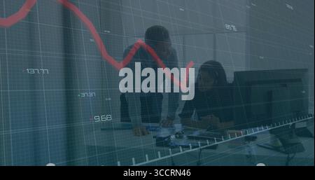 Collaborating two professionals writing documents at office desk, with monitors  and  red chart over Stock Photo