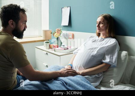 Caucasian pregnant woman lying in hospital bed holding belly while middle aged Caucasian man sitting beside her touching hand, both focusing on each other in medical setting Stock Photo