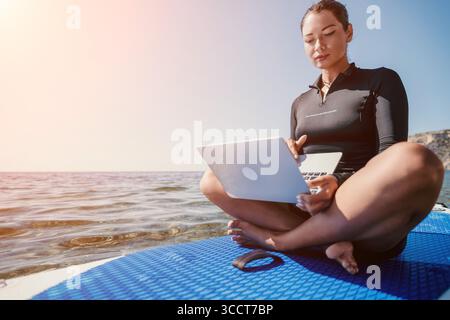 Woman Laptop Beach Work Remotely - Young woman uses laptop on paddleboard while enjoying the beach on a sunny day. Stock Photo