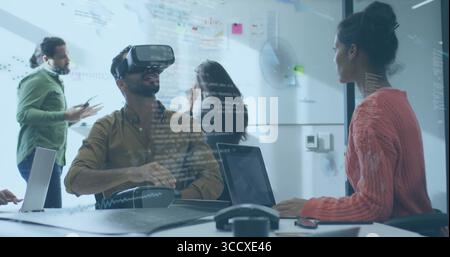 VR headset wearer in brown shirt using control pad in office meeting, laptops, whiteboard notes Stock Photo