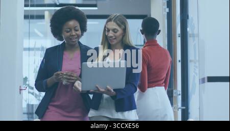 Two women walking and examining laptop in corridor with frosted partitions, holding glass of water Stock Photo