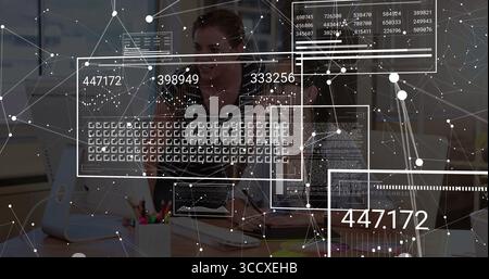 Adult Asian analyst analyzing data overlays at desk in office, with dual monitors and keyboard Stock Photo